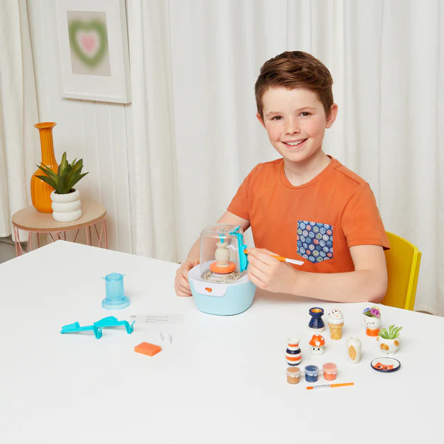 Boy sitting at table using mini pottery wheel with completed pieces of pottery to the side