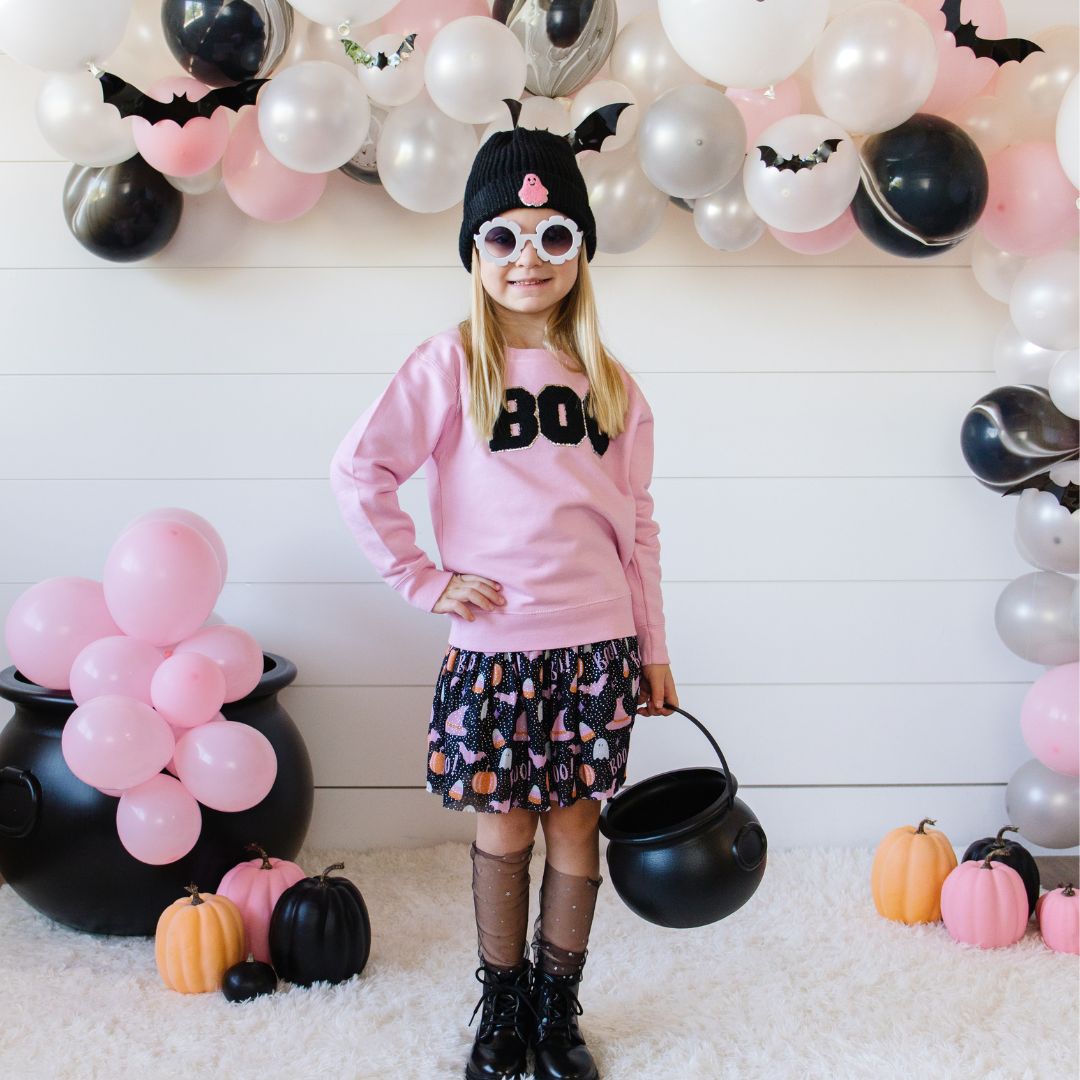 Girl with pink sweatshirt with black BOO patch in front of balloon arch full of silver, white, pink and black balloons